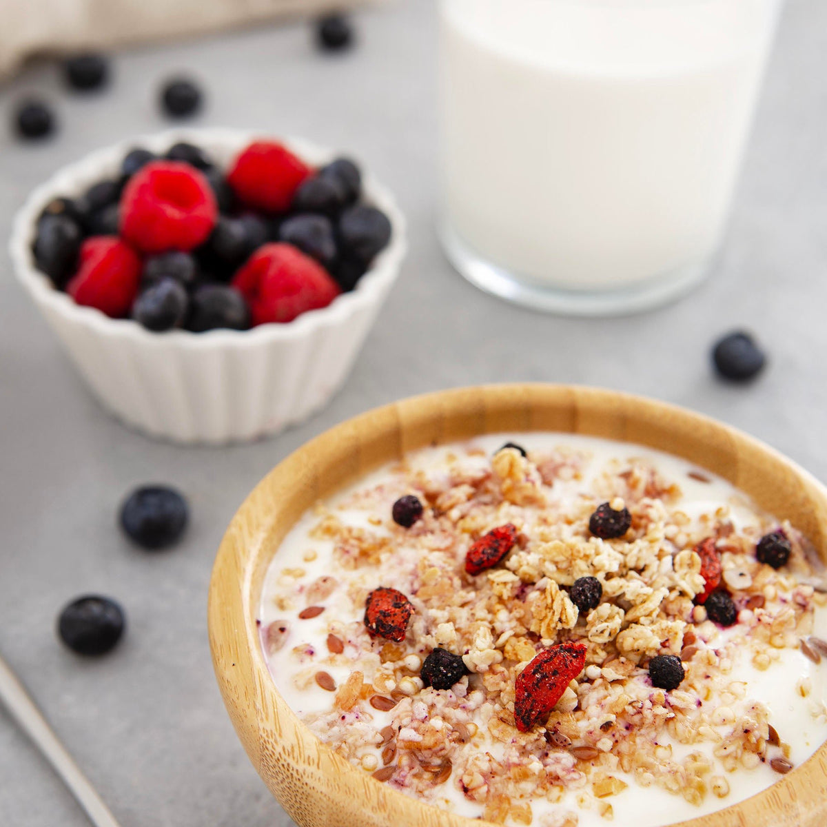 Ciotola con yogurt, granella e frutti rossi, con lamponi e mirtilli sullo sfondo.