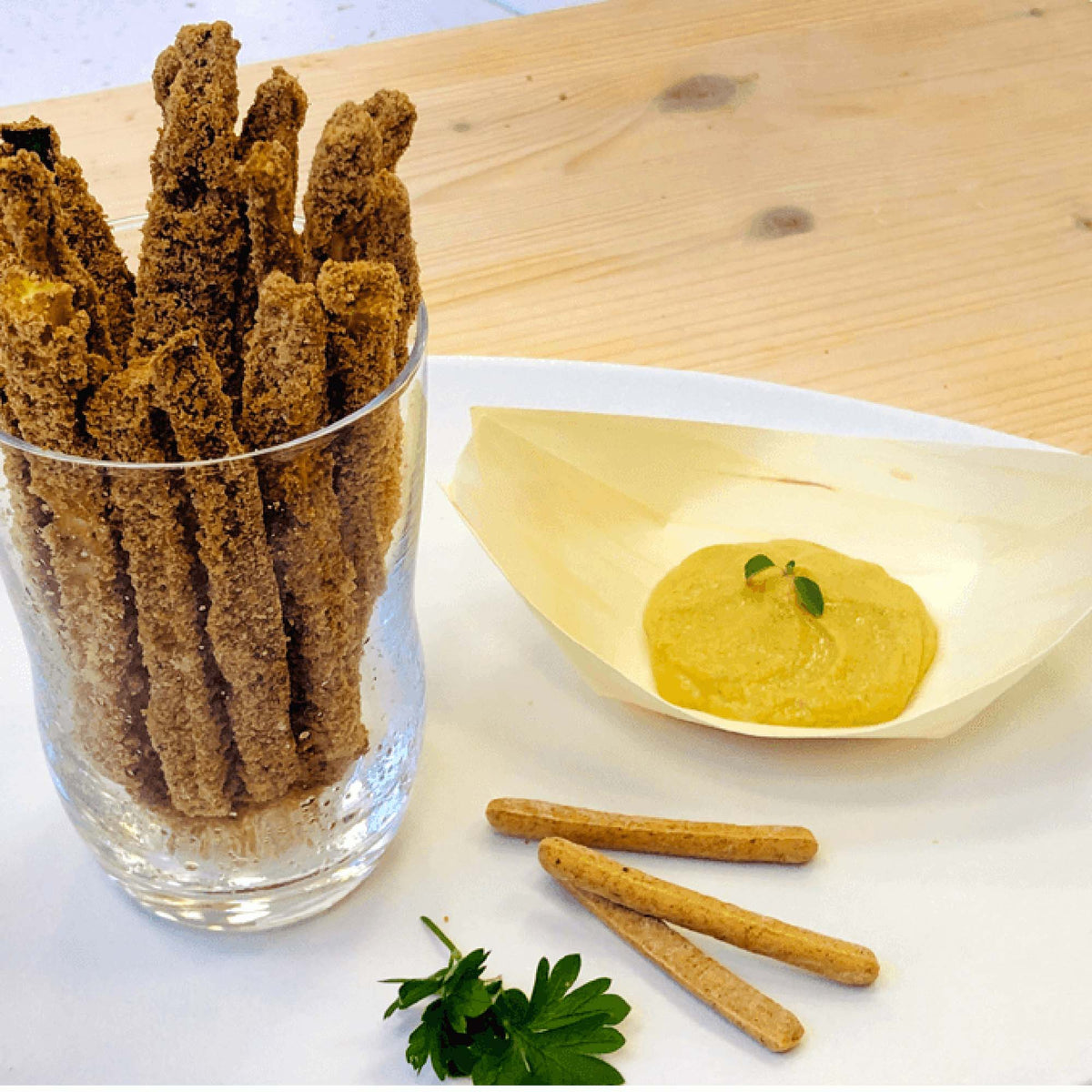Grissini ricoperti da una panatura scura in un bicchiere, accanto a una crema gialla e grissini al naturale.