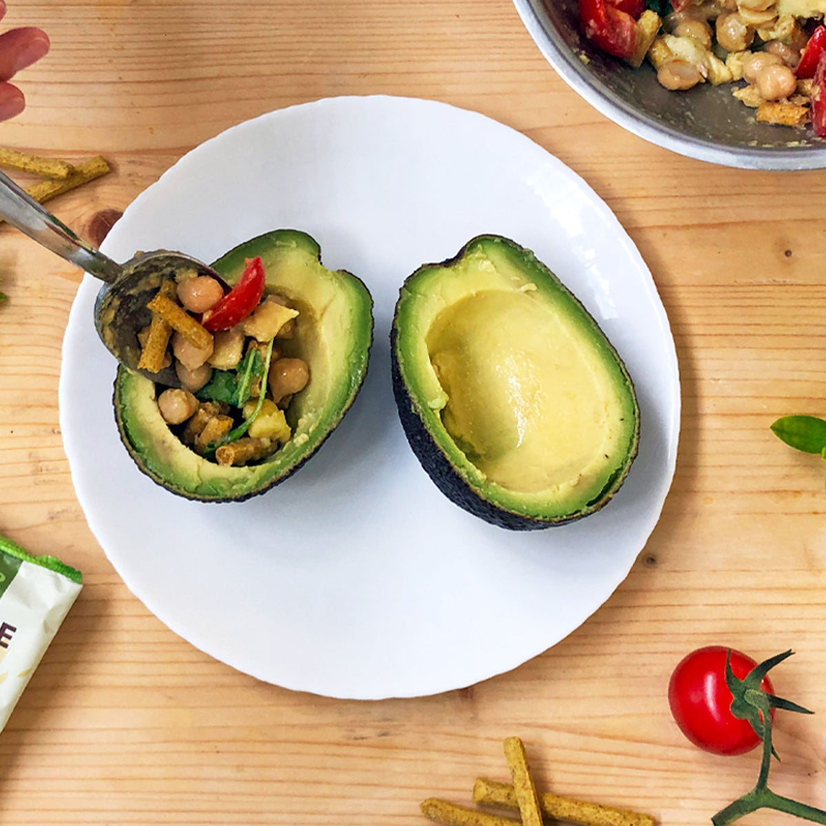 Metà di avocado farcita con ceci, pomodorini e pezzi di grissini, su un piatto bianco.